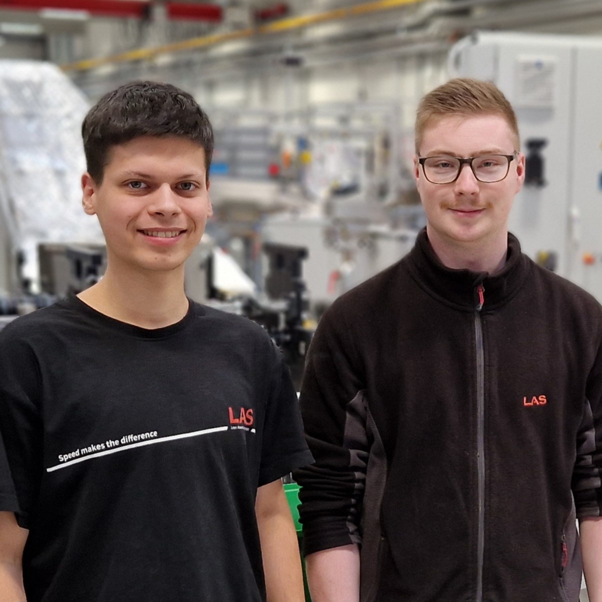Die ausgebildeten Mechatroniker Joel Nowak und Timo Werner stehen vor einer Montageanlage in der LAS-Montagehalle.